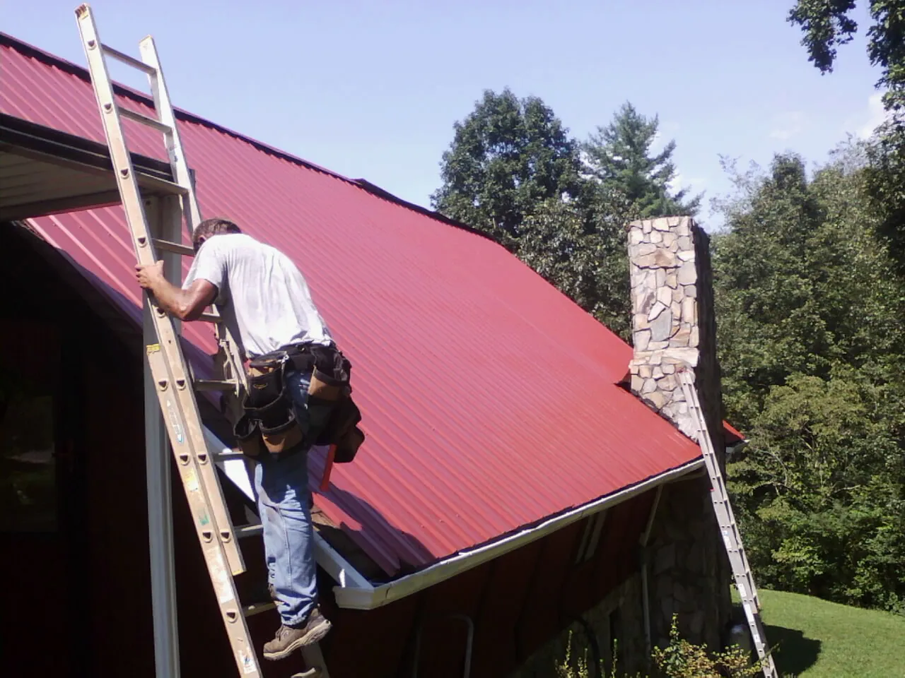 Roofer on a ladder beside a red metal roof and stone chimney — Soffit & Fascia Repair by Roofing Brothers in Iowa Falls